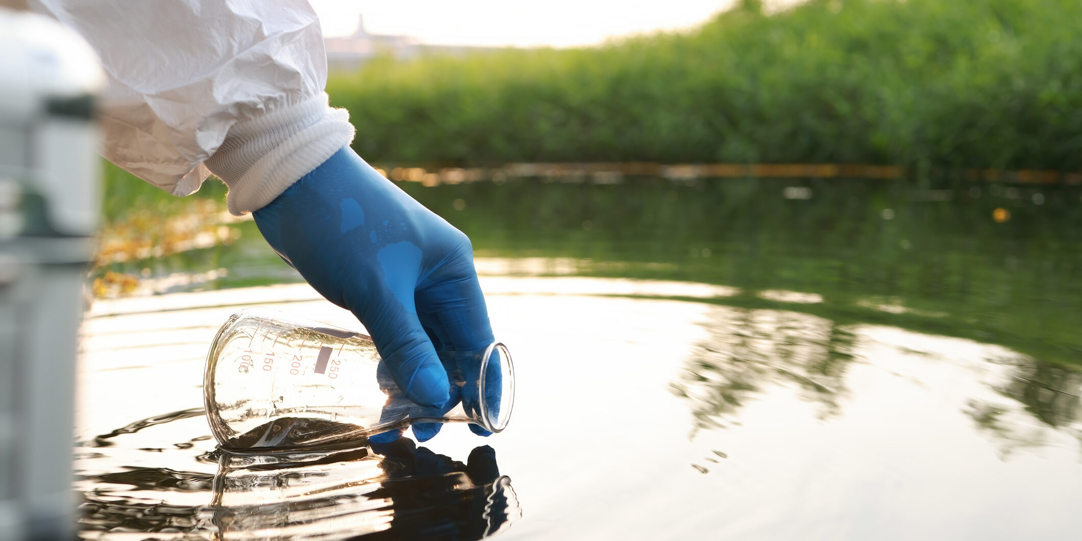 Close up hand with glove Collecting samples of water with a test tube from a body of water with trees in the background