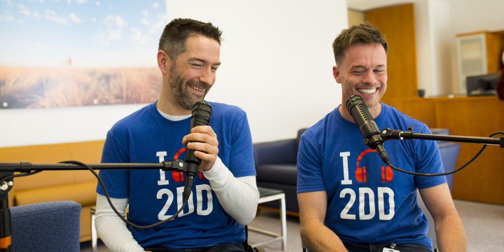 Kyle Bryant and Sean Baumstark laugh behind two microphones. Kyle is a white man with dark brown hair and a blue T-shirt that says "I (image of headphones) 2DD." Sean is a white man with blonde hair and is wearing the same T-shirt as Kyle.
