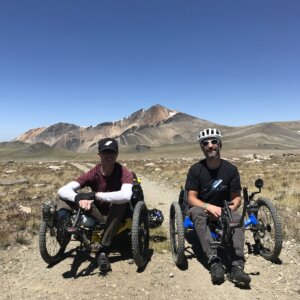 Sean and Kyle wear helmets and sunglasses as they sit on racing trikes on a mountain path.
