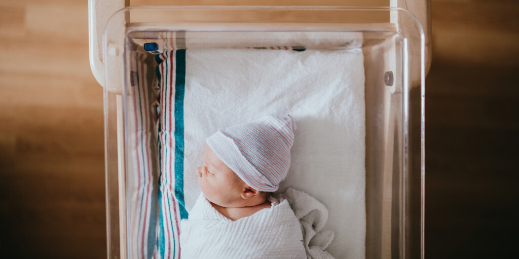 A baby just born at the hospital rests in a hospital bassinet crib, wrapped in a swaddle and wearing a beanie hat.