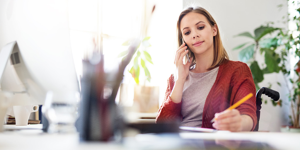 A woman in a wheelchair sits behind a desk holding a telephone to her ear and a pencil in her other hand