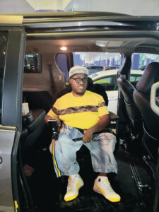 Miguel Morris sits inside a van in a power wheelchair. He is wearing a bright yellow shirt, light blue jeans, and white sneakers.