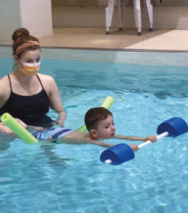 Four-year-old William Shirk floats on his stomach in a swimming pool with an aquatic therapist standing behind him in the pool.