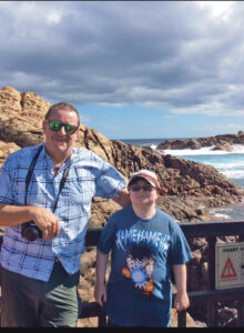 Alt TextResearcher Steve Wilton, PhD, BSc (Hons), wearing casual clothes and carrying a camera, stands next to young clinical trial participant Billy Ellsworth. Behind them is a rocky coastline.