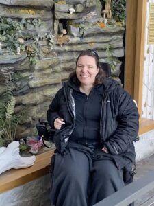 Crystal Killian, dressed in a black down jacket, sits in a power wheelchair in front of a rock wall growing ferns and ivy.