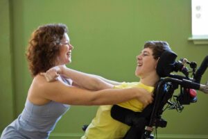 Yoga teacher Kerri Hanlon leans toward a young man seated in a power wheelchair. They both have their arms extended to touch each other’s shoulders.