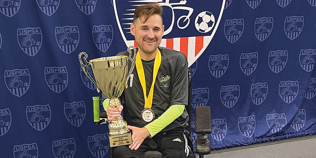 Chad Wilson sits in a power wheelchair. He is wearing a medal and holding a trophy.