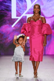 A young girl in a silver fringe dress waves while walking with a woman in a pink dress on a fashion runway