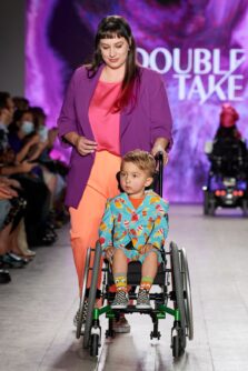 A young boy in a wheelchair wears a popsicle print suit on a fashion runway while his mother walks beside him in a purple jacket
