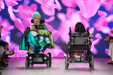 A man in a wheelchair rolls on the runway of a fashion show wearing a colorful cape and hat while another model in a wheelchair rolls away from the camera