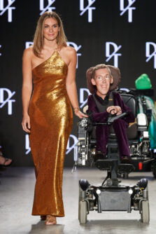 A woman in a gold, one shoulder gown walks the runway next to a man in a velvet suit using a wheelchair