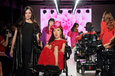 A woman in a red dress and red hat uses a powerchair on a fashion runway, a woman in a black dress walks beside her, and a model in a wheelchair rolls away from the camera