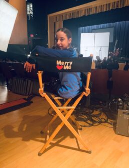 A teenage girl sits in a directors chair on set of the film "Marry Me" and the title of the movie can be seen on the back of the chair while she twists to smile at the camera