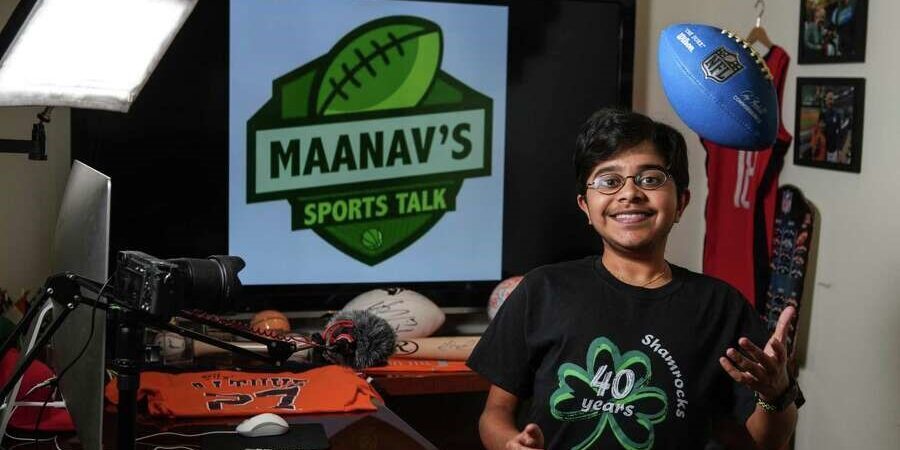Maanav Gupta stands in front of sign that reads Maanav's Sports Talk surrounded by recording and sports equipment