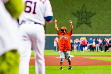 Maanav on baseball field in Astros jersey with both hands thrown in the air to celebrate throwing first pitch of the season