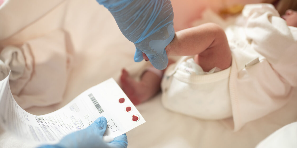 Hands with rubber gloves hold the heel of an infant in one hand and a piece of paper with newborn testing sample in the other hand.