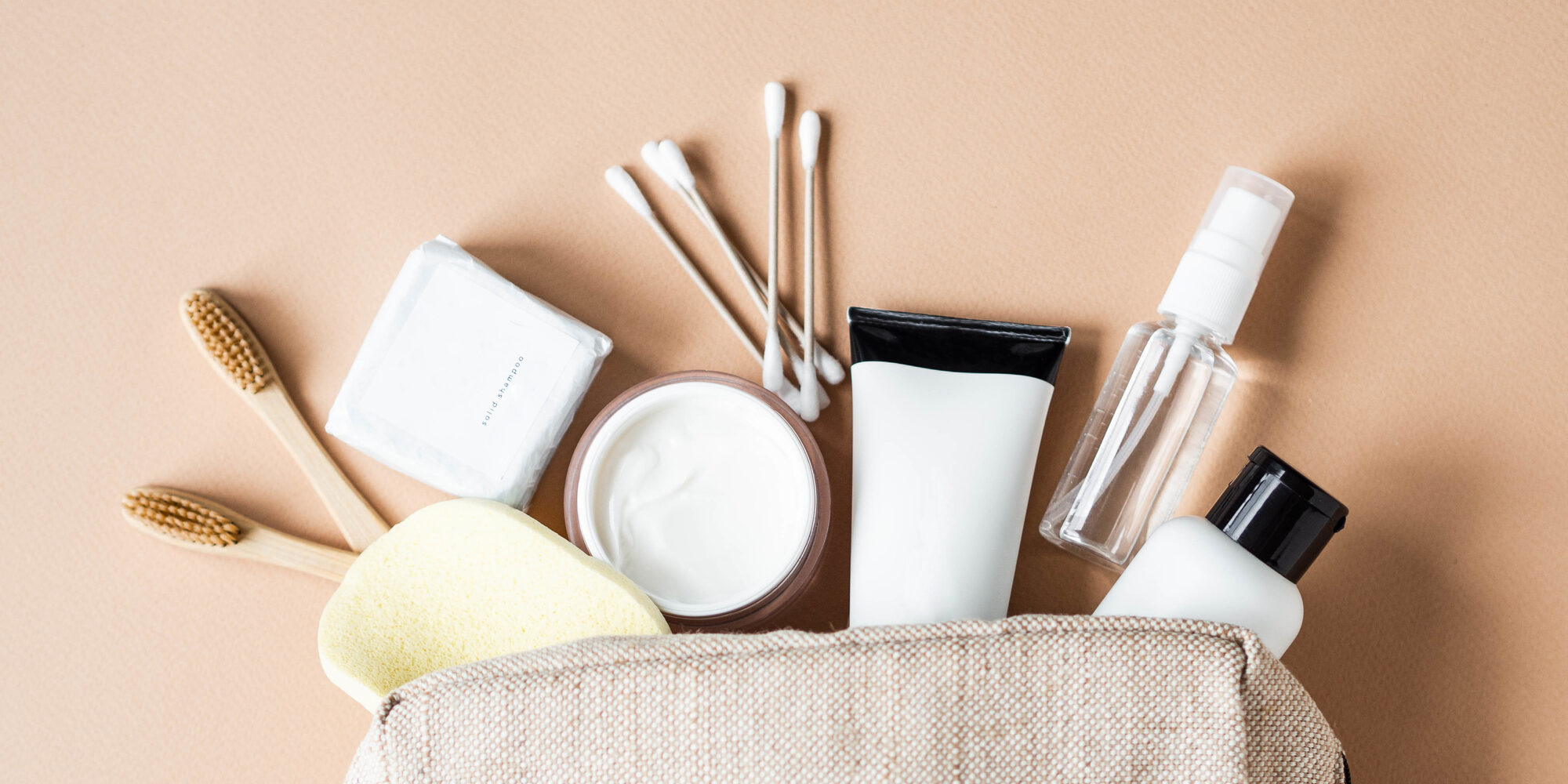Hygiene products, dry shampoo, cotton buds, toothbrushes poking out of bag on a beige background.