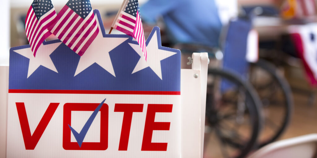 Vote sign with three small America flags and a partial view of a man in wheelchair in the background