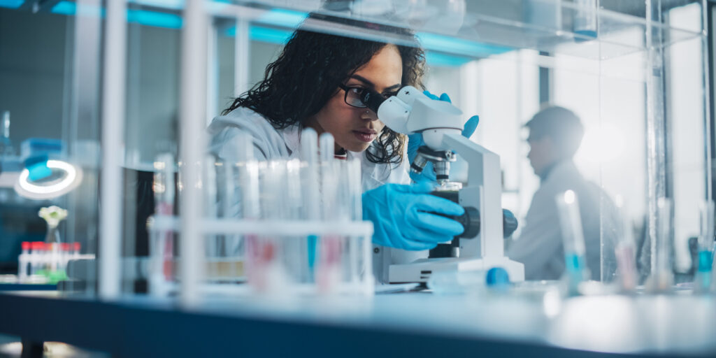 Female Scientist Looking Under Microscope with test tubes on her table