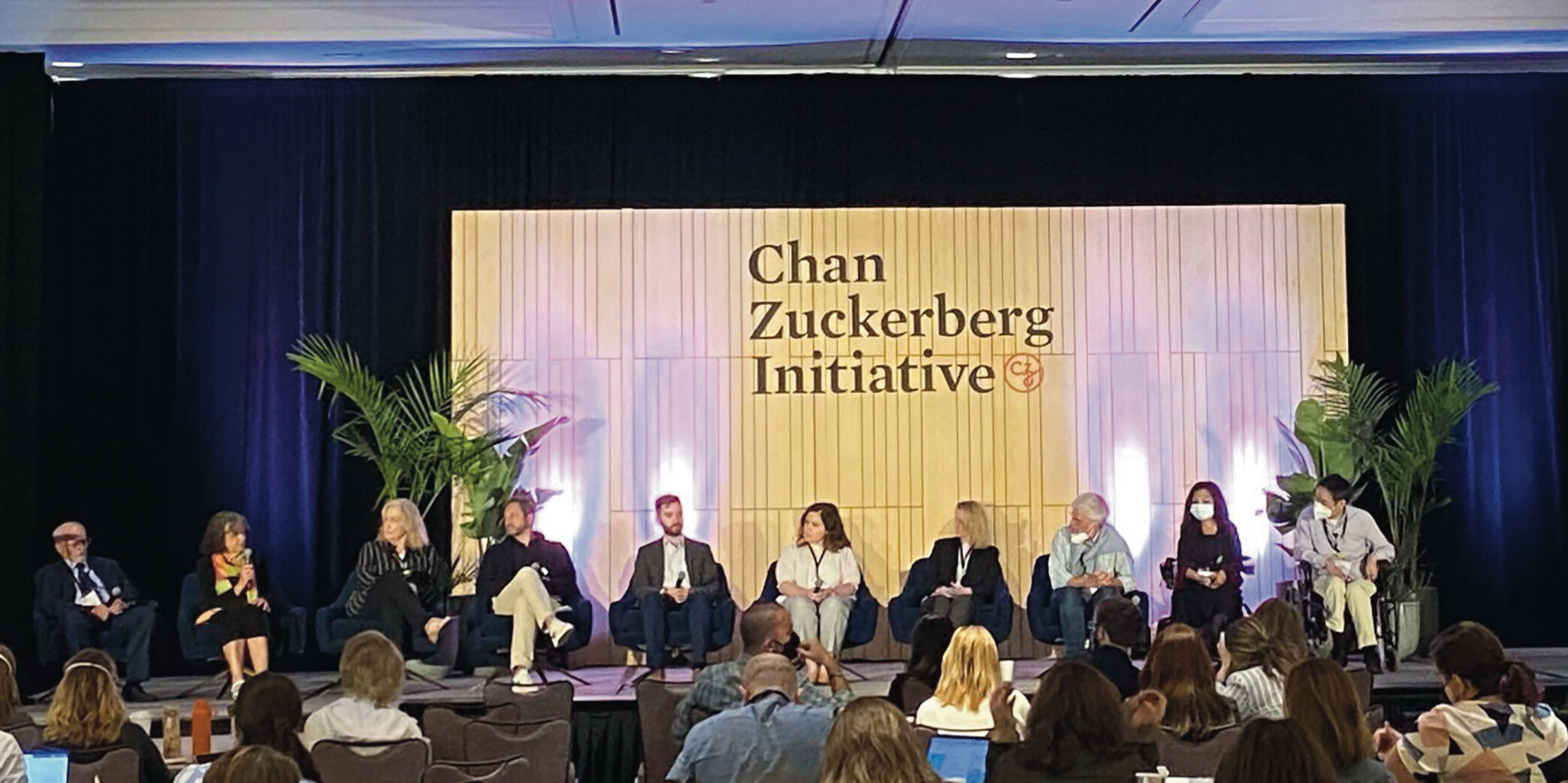 A panel of adult males and females at the Chan Zuckerberg Initiative conference sitting down engaged in a discussion on stage. Two individuals are seated in wheelchairs, several are wearing face masks and holding microphones.