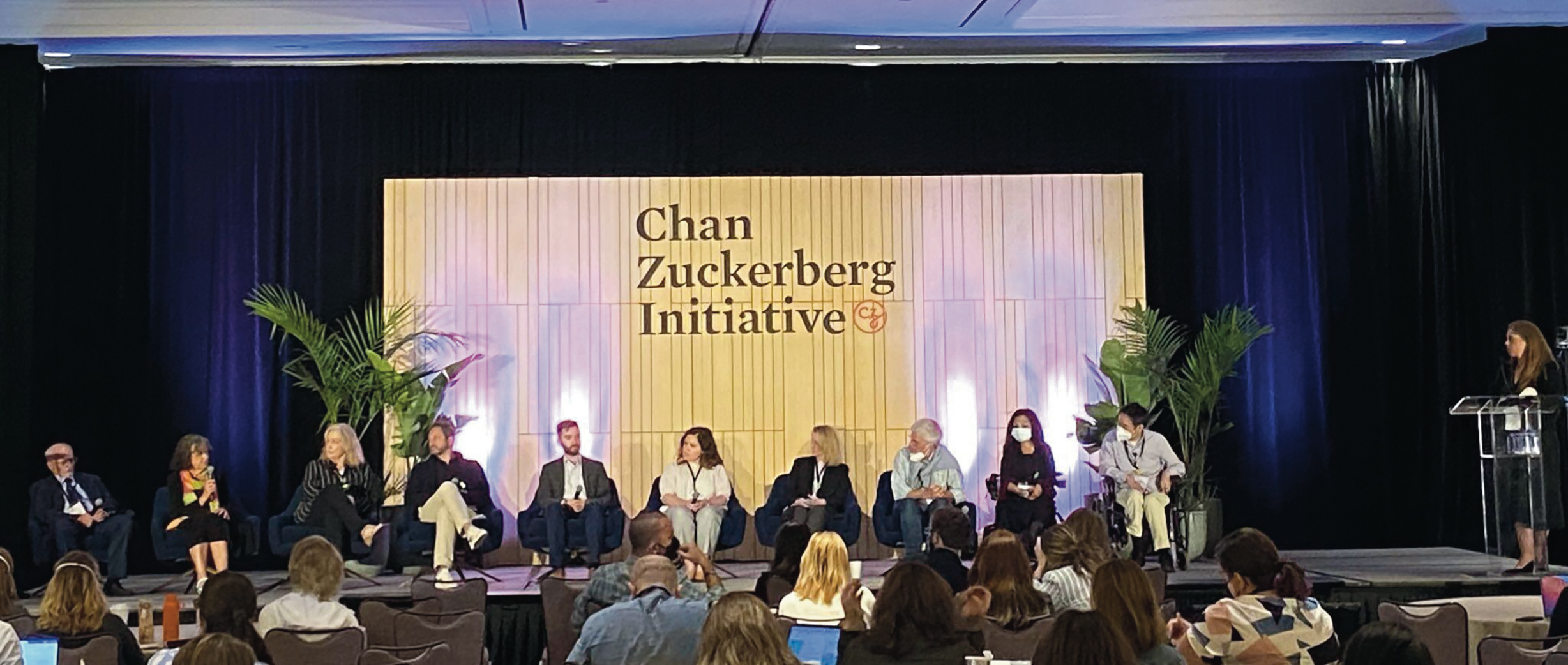 A panel of adult males and females at the Chan Zuckerberg Initiative conference sitting down engaged in a discussion on stage. Two individuals are seated in wheelchairs, several are wearing face masks and holding microphones.