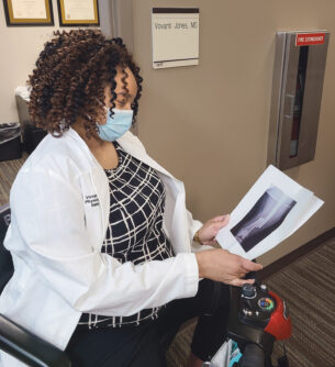 Dr. Vovanti Jones, a Black woman with curly brown hair wearing a white lab coat and surgical mask, is looking down at an X-ray of a broken bone she holds in her hands. She is sitting in a motorized scooter in front of an office labeled with her name.