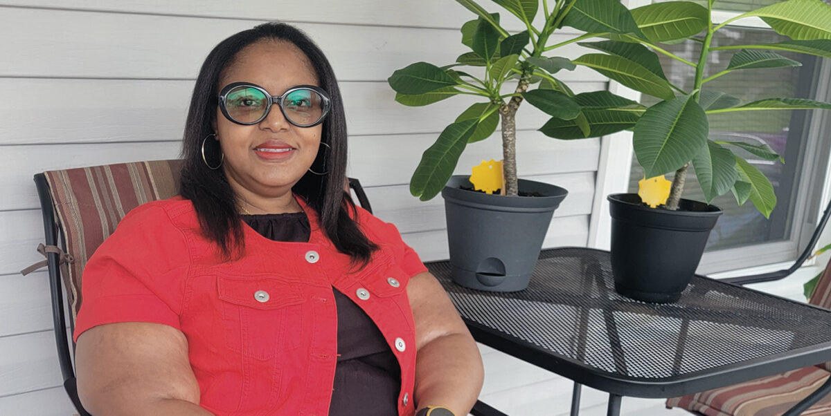 Tomeca Goodwin, a Black woman, is sitting outdoors in a patio chair smiling, next to two potted plants. She’s wearing a red short-sleeve cardigan, long black dress, and strappy red sandals. She also sports round, black-rimmed glasses and gold hoop earrings.