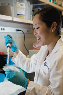 Dr. Angela Lek, an Asian woman with black hair pulled into a bun, wearing a white lab coat and blue rubber gloves, prepares a research sample using a pipette and testing plate.