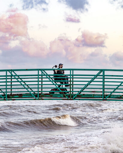 Photo of man in power wheelchair on bridge over ocean waves and sky with clouds