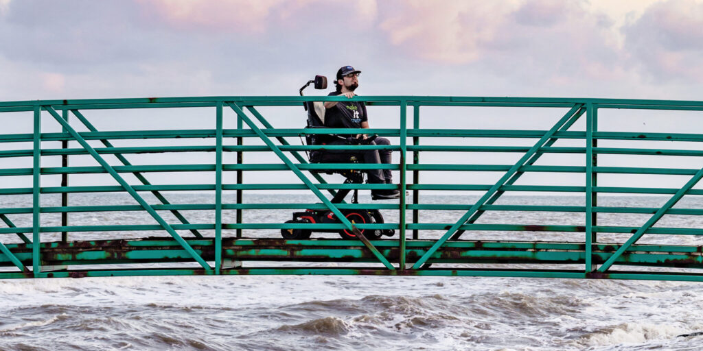 Sean Wallace, a young white man with a black baseball cap, dark brown beard, and black shirt and pants, sits in a black power wheelchair in the center of a gently arching bridge with green railings. Neither end of the bridge is visible in the photo, so the man and bridge appear to be suspended in a sky with purple-tinged clouds above white-capped sea waves.