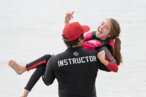 A man in a red cap and black wetsuit labeled “instructor,” seen from behind, carries Abby, a white girl with brown hair in a ponytail, toward a body of water. Abby, wearing a black and red wetsuit and life jacket, is looking over his shoulder, smiling and laughing.