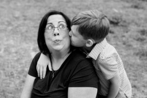 Black and white close-up photo of Susan Manning, a white woman with dark, chin-length hair, looking up with a surprised expression on her face while her young son, a boy with light hair and a striped shirt, kisses her on the cheek.