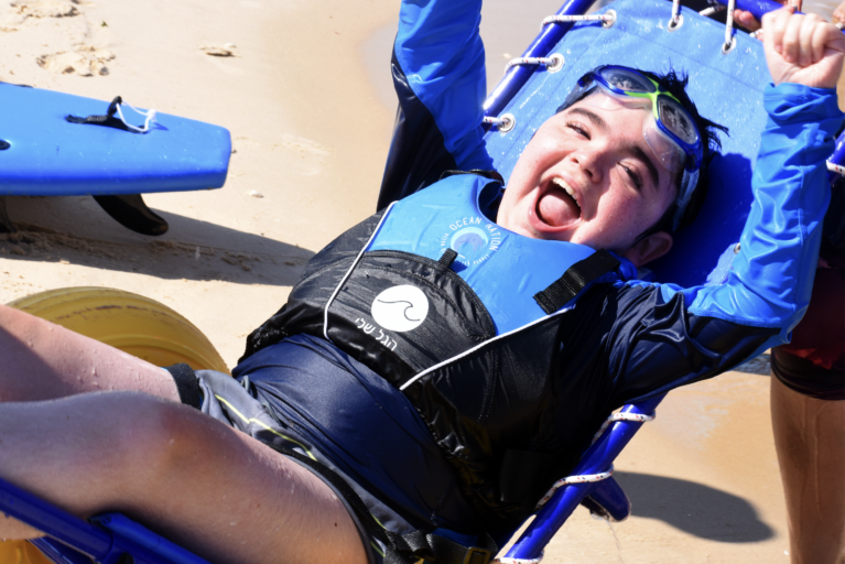 A boy with light skin and dark hair and swim goggles perched on his forehead looks at the camera with a large, open-mouthed smile and his hands up in the air. He wears a blue long-sleeve rash guard and life vest and sits in a beach wheelchair with large, yellow inflated wheels designed to roll on sand.