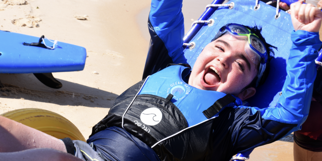 A boy with light skin and dark hair and swim goggles perched on his forehead looks at the camera with a large, open-mouthed smile and his hands up in the air. He wears a blue long-sleeve rash guard and life vest and sits in a beach wheelchair with large, yellow inflated wheels designed to roll on sand.