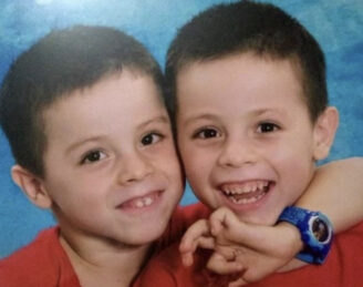 Close up of Jean Carlos and Jeandy Daniele, identical twins with light skin and dark brown hair and eyes, embracing.
