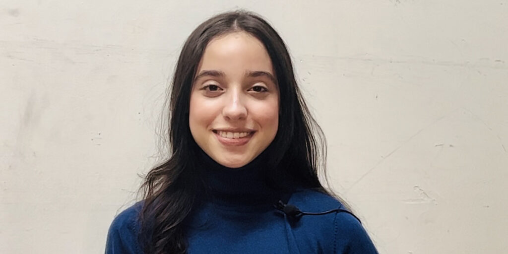 Headshot of Leah Zelaya, a teenage girl with long brown hair wearing a blue turtle neck against a tan background