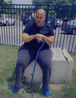 Quanetta Holt, a Black woman with a smooth-shaven head wearing a black T-shirt and dark pants, sits on a stone bench with her eyes closed and her hands resting on a blue walking cane.