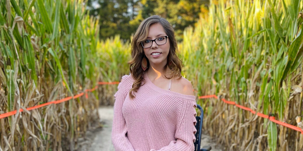 Skye Anderson, a white woman with long, wavy brown hair, brown-rimmed glasses, dangly earrings, and a pink sweater off one shoulder, sits in a wheelchair on a path between rows of tall green and brown cornstalks.