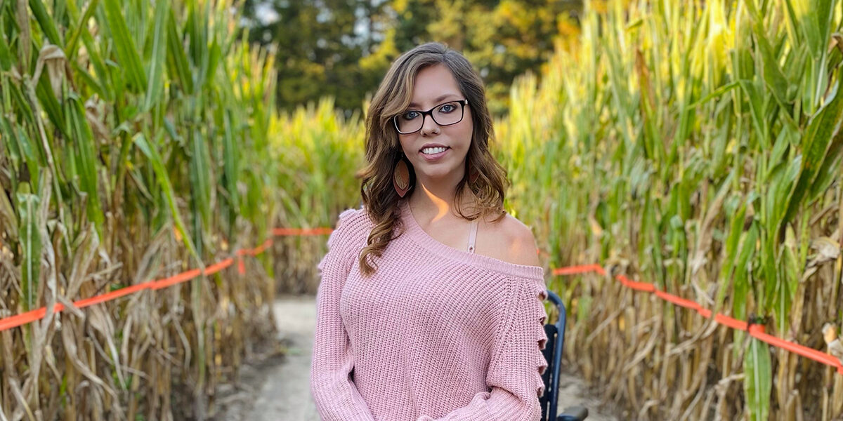 Skye Anderson, a white woman with long, wavy brown hair, brown-rimmed glasses, dangly earrings, and a pink sweater off one shoulder, sits in a wheelchair on a path between rows of tall green and brown cornstalks.