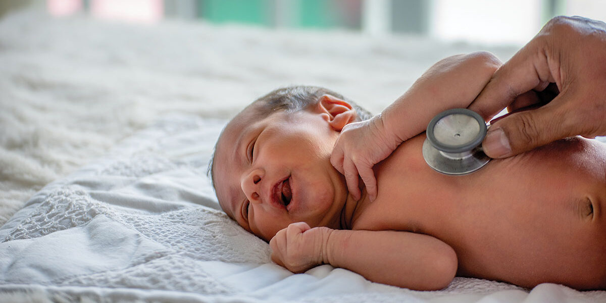 Closeup of an unclothed infant lying on its side on a white blanket while an adult hand holds a stethoscope to its chest.