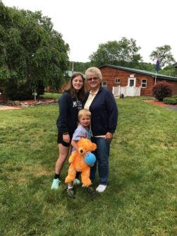 Volunteer counselor, a camper holding a large teddy bear, and the campers grandma stand outside of a camp cabin.