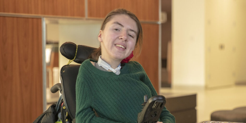 Katie Frayer smiles while wearing a green sweater and sitting in her power chair