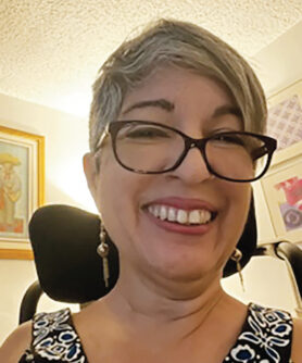 Closeup of a smiling middle-aged Caucasian woman with short gray hair, black-rimmed glasses, and dangly earrings with silver balls and tassels.