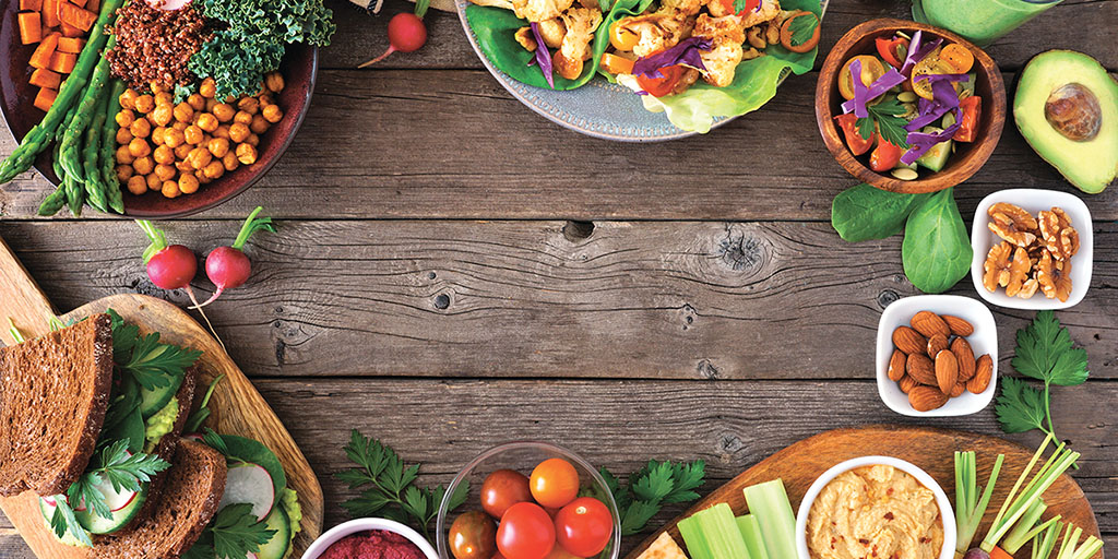 A brown wooden table with a collection of bowls and plates holding healthy foods, including tomatoes, chickpeas, radishes, leafy greens, nuts, and avocado.