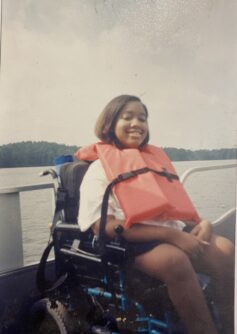 A young girl in a wheelchair wearing a life vest sits in front of a lake