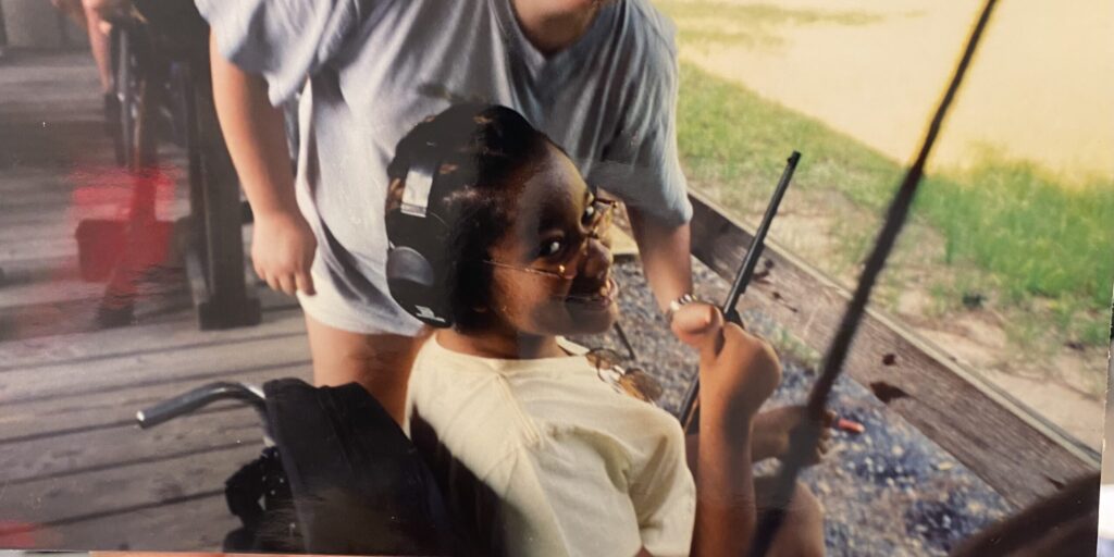 A young Leticia smiles from her wheelchair holding a fishing rod at MDA Summer Camp