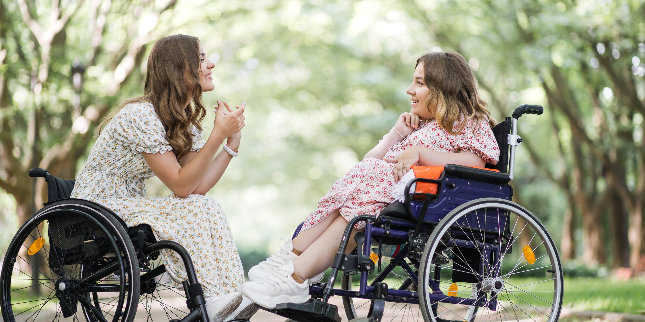 Side view of two happy women in wheelchairs talking and smiling outdoors. Background of green summer park.