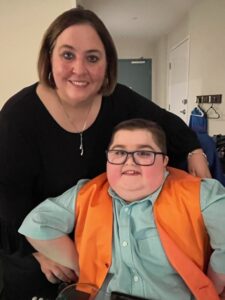 Christy Hickman, a white woman with chin-length straight, brown hair stands behind her son Peyton, a white boy with short brown hair and black-rimmed glasses, wearing a green button-down shirt and orange vest.