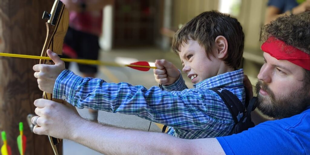 Side view of volunteer Devin Jensen helping a camper hold a bow and arrow