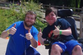 Devin wears a blue t-shirt and gives a double thumbs up next to a camper in a wheelchair also giving thumbs up